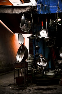 An assortment of metal kitchenware is displayed outdoors, including large pots, pans, and strainers, hanging and stacked against a dimly lit backdrop. A blue tarp covers part of the setup, and an orange wall provides a warm glow to the left.