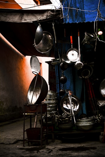 An assortment of metal kitchenware is displayed outdoors, including large pots, pans, and strainers, hanging and stacked against a dimly lit backdrop. A blue tarp covers part of the setup, and an orange wall provides a warm glow to the left.