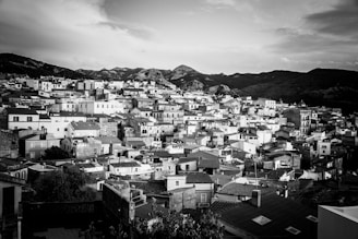 A densely packed array of buildings with uniform, rectangular shapes, all in monochrome, stretches across a hillside. The background features a range of hills or small mountains under a cloudy sky, creating a striking contrast with the urban landscape.