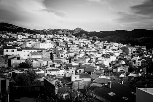 A densely packed array of buildings with uniform, rectangular shapes, all in monochrome, stretches across a hillside. The background features a range of hills or small mountains under a cloudy sky, creating a striking contrast with the urban landscape.