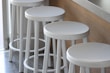 Four white bar stools are aligned in a row next to a wooden counter. The setting appears to be modern and minimalistic, providing a clean and organized aesthetic. The stools have a sleek design, and the overall arrangement suggests a casual and inviting atmosphere.