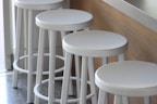 Four white bar stools are aligned in a row next to a wooden counter. The setting appears to be modern and minimalistic, providing a clean and organized aesthetic. The stools have a sleek design, and the overall arrangement suggests a casual and inviting atmosphere.
