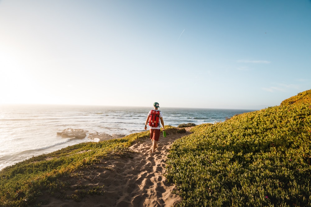 man walking while carrying red backpack near sea at daytime