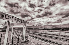 A black and white image of a rustic railway track with a sign reading 'Klaas Voogdsrivier' in the foreground. The setting appears to be a rural, mountainous area under a cloudy sky, conveying a sense of abandonment or solitude.