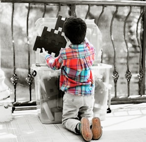 A child wearing a colorful plaid shirt and khaki pants is kneeling on the floor, interacting with large, interlocking black puzzle pieces stored in a clear plastic container. The setting appears to be an indoor or semi-outdoor space with a decorative railing in the background.