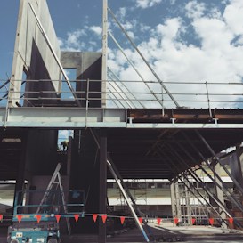 A construction site with metal scaffolding and partially built structures against a backdrop of a blue sky with scattered clouds. There are ladders and construction equipment, including a lift, visible on the ground. Red warning flags are strung across the foreground.