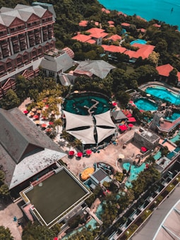 An aerial view of a resort complex with multiple buildings featuring red roofs surrounded by lush greenery. Several swimming pools with turquoise water are visible, alongside poolside areas with red umbrellas and lounge chairs. A large canopy structure is centrally located, and there are walking paths and landscaped gardens throughout the area.