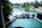 A luxurious resort setting with a large, inviting swimming pool surrounded by palm trees. There is a wooden deck area with dining tables and chairs arranged in a neat fashion. The atmosphere is serene and tropical, with the pool reflecting the greenery of the surrounding trees.