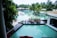 A luxurious resort setting with a large, inviting swimming pool surrounded by palm trees. There is a wooden deck area with dining tables and chairs arranged in a neat fashion. The atmosphere is serene and tropical, with the pool reflecting the greenery of the surrounding trees.