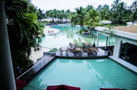 A luxurious resort setting with a large, inviting swimming pool surrounded by palm trees. There is a wooden deck area with dining tables and chairs arranged in a neat fashion. The atmosphere is serene and tropical, with the pool reflecting the greenery of the surrounding trees.
