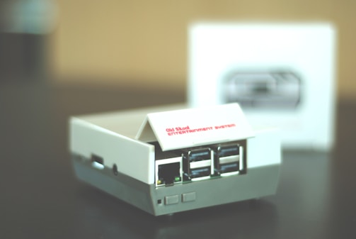 A small retro gaming console designed to resemble an old school entertainment system, featuring multiple USB ports on its front. The console is primarily white with some gray accents, and it sits on a dark surface. A blurred box with an image of the console is visible in the background.