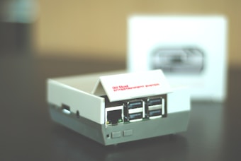 A small retro gaming console designed to resemble an old school entertainment system, featuring multiple USB ports on its front. The console is primarily white with some gray accents, and it sits on a dark surface. A blurred box with an image of the console is visible in the background.