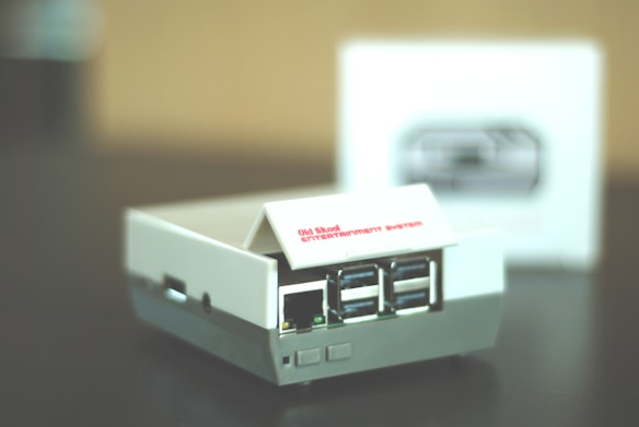 A small retro gaming console designed to resemble an old school entertainment system, featuring multiple USB ports on its front. The console is primarily white with some gray accents, and it sits on a dark surface. A blurred box with an image of the console is visible in the background.