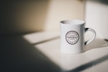 A white ceramic coffee mug with a circular logo bearing the text 'COOPER & CO, COFFEE ROASTERS, ST HELIER, JERSEY, TEA MERCHANTS' is positioned on a flat surface. The mug is partially illuminated by soft, natural light coming from an unseen source, creating gentle shadows.