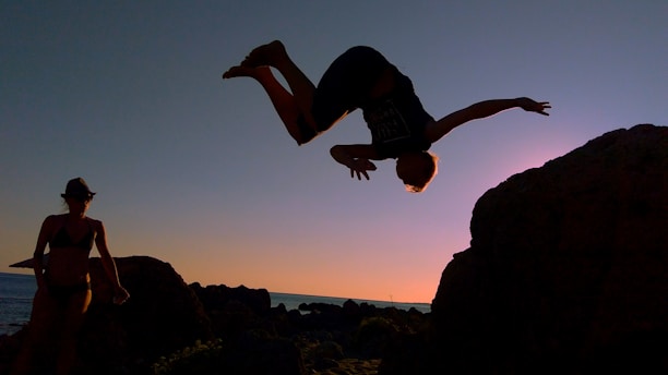 A person is performing a backflip off a rocky surface against a sunset backdrop, creating a silhouette against the sky. Another person stands nearby, watching the action.