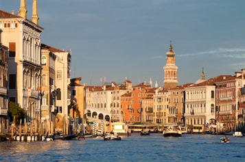 A picturesque view of a canal with water taxis and gondolas moving along it. The buildings lining the canal are a mix of historic architecture with ornate facades and soft pastel colors. A bell tower stands prominently in the background under a clear blue sky.