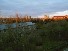 A residential area with trees in the foreground and several buildings in the background, illuminated by the warm light of a sunset or sunrise. The sky is mostly cloudy, casting a soft light over the scene.
