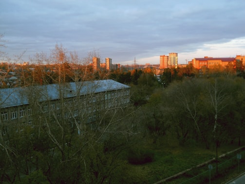 A residential area with trees in the foreground and several buildings in the background, illuminated by the warm light of a sunset or sunrise. The sky is mostly cloudy, casting a soft light over the scene.