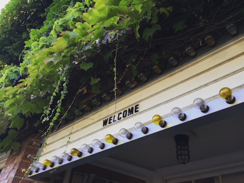 A warm, sunlit farmhouse porch with blooming flowers and a rustic wooden sign that reads 'Welcome to Alexa Farms.'