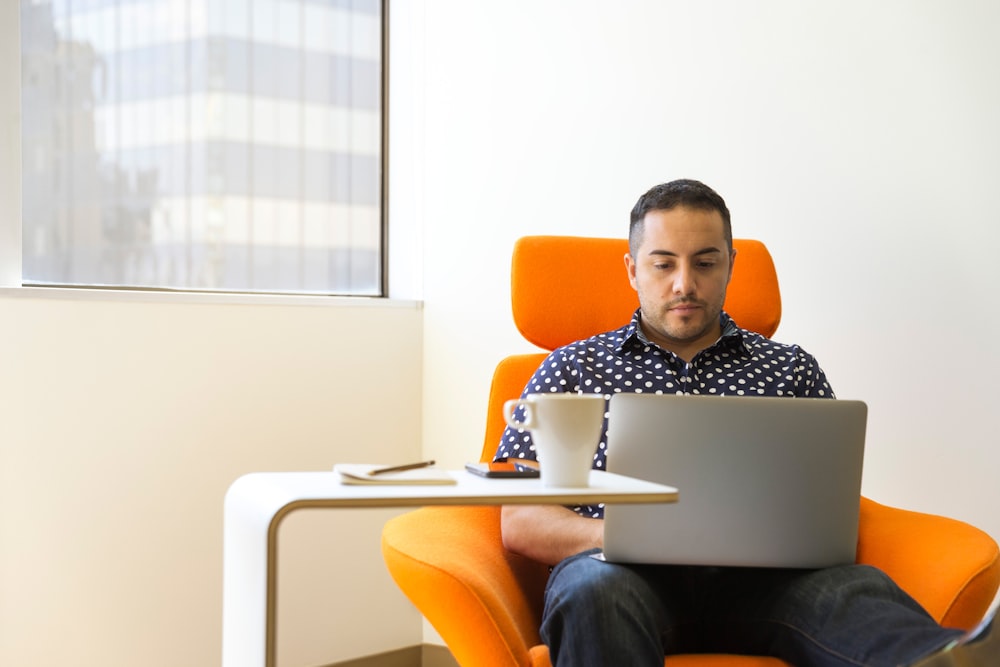 homem vestindo camisa de gola de bolinhas azul e branca sentado na cadeira do sofá de tecido laranja enquanto usa laptop prateado ao lado da mesa de madeira branca com caneca de cerâmica branca