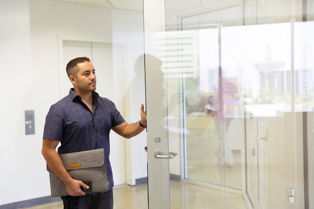 hombre con cuello azul abotonado en la parte superior que sostiene la bolsa gris de la computadora portátil y sostiene la puerta de vidrio