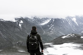 A person wearing a black backpack and dark clothing stands on a rocky landscape, gazing at a mountainous scene with snow patches and misty clouds covering the mountain peaks.
