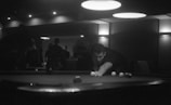 A dimly lit room with several people focused around a pool table. A man is preparing to take a shot, leaning over to aim at the cue ball. Overhead lights create a moody atmosphere, and silhouettes of other people can be seen in the background.