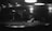 A dimly lit room with several people focused around a pool table. A man is preparing to take a shot, leaning over to aim at the cue ball. Overhead lights create a moody atmosphere, and silhouettes of other people can be seen in the background.