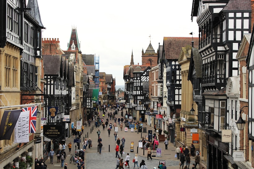 A bustling street is lined with historic black-and-white Tudor-style buildings featuring ornate details. People are walking along the street, some stopping to look at shops or signs. Various shop signs and banners are visible, including a prominent Union Jack flag. The architecture features steeply pitched roofs and intricate woodwork, typical of English heritage. In the background, church spires and red brick structures add to the historic atmosphere.