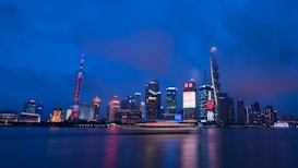 The image captures a vibrant city skyline at night, with skyscrapers illuminated by colorful lights and a tall tower featuring unique architecture. Bright neon signs and the reflection of the cityscape on a river add to the dynamic scene.