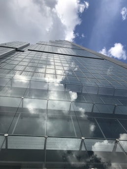 A tall, modern skyscraper with reflective glass windows stretches upwards towards a partly cloudy blue sky. The reflection of clouds on the windows creates a seamless blend between the structure and the sky.