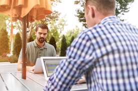 Two men are working on laptops at an outdoor patio table under an orange umbrella. One man is facing the camera, wearing a green shirt, while the other is in a blue checkered shirt with his back to the camera. The setting is surrounded by greenery, creating a serene and productive atmosphere.