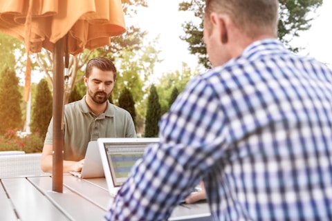 Two men are working on laptops at an outdoor patio table under an orange umbrella. One man is facing the camera, wearing a green shirt, while the other is in a blue checkered shirt with his back to the camera. The setting is surrounded by greenery, creating a serene and productive atmosphere.