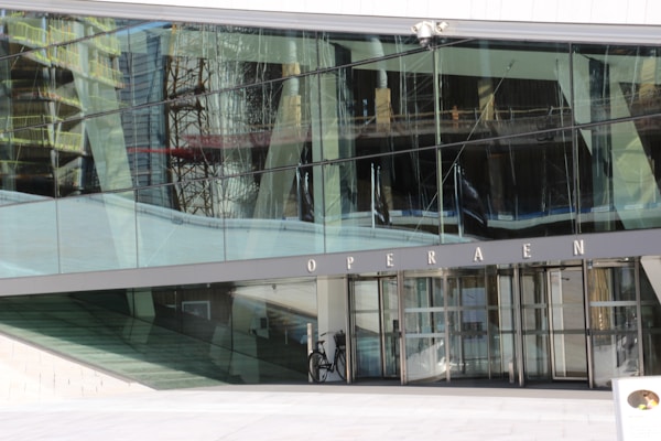 A modern building with large reflective glass panels that show distorted reflections of another structure with scaffolding. The entrance is marked by the word 'OPERAEN' above a set of glass doors. A bicycle is parked near the entrance, and the scene appears quiet and unoccupied.