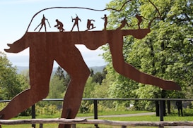 A large rust-colored metal sculpture depicting the evolution of humans is set against a backdrop of lush green trees. The figures on the sculpture appear in various stages of evolution, from an early hominid to a modern human. The scene is bright and natural, with the metal artwork sharply contrasting with the vivid greenery.