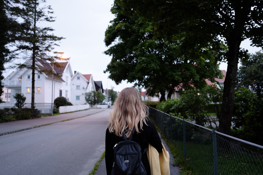 woman wearing black long-sleeved shirt and backpack walking on gray concrete road between green trees near white concrete house during daytime