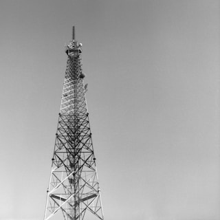 A tall, metallic radio or communication tower set against a clear sky. The structure consists of a lattice framework, tapering as it ascends, with various antennas attached.