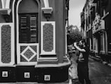 A person holding a camera stands in a narrow street lined with buildings featuring ornate architectural details. The scene is captured in black and white, highlighting the textures and patterns of the walls and windows.