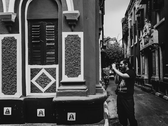 A person holding a camera stands in a narrow street lined with buildings featuring ornate architectural details. The scene is captured in black and white, highlighting the textures and patterns of the walls and windows.