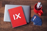 A red notebook with a white 'IX' logo is placed on top of a closed laptop on a wooden table. Nearby, a red mug holds several pens and markers. A stack of business cards with a blue background and the 'imgIX' logo lies next to the mug.