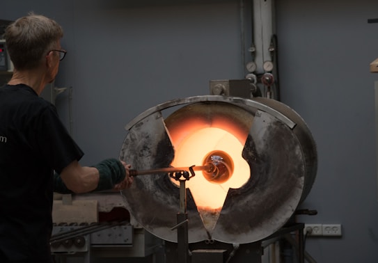 A person engaged in glassblowing is holding a rod with molten glass in front of a glowing furnace. The intense orange light from the furnace contrasts with the dark surroundings, creating a dramatic effect. The individual is wearing protective gear and appears focused on shaping the glass piece.