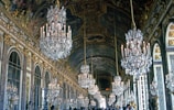 An opulent hall adorned with large, intricate crystal chandeliers suspended from the ornate ceiling. The walls are lined with mirrors and decorated with gold accents and artistic details. A crowd of people is visible, reflecting the grandeur and historic significance of the location.