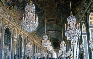 An opulent hall adorned with large, intricate crystal chandeliers suspended from the ornate ceiling. The walls are lined with mirrors and decorated with gold accents and artistic details. A crowd of people is visible, reflecting the grandeur and historic significance of the location.