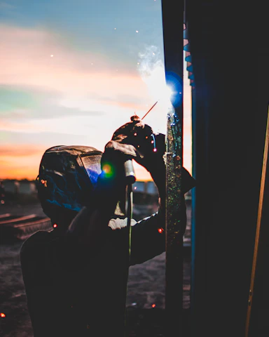 A skilled welder in protective gear performing aluminum welding on a truck frame at dusk.