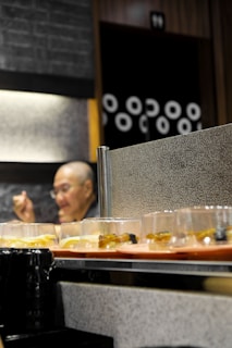 A conveyor belt with various plates of sushi passes by. In the background, a person appears to be sitting and possibly eating. The wall behind them is adorned with a pattern of circles.