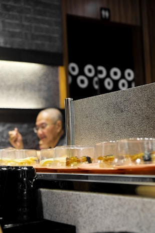 A conveyor belt with various plates of sushi passes by. In the background, a person appears to be sitting and possibly eating. The wall behind them is adorned with a pattern of circles.