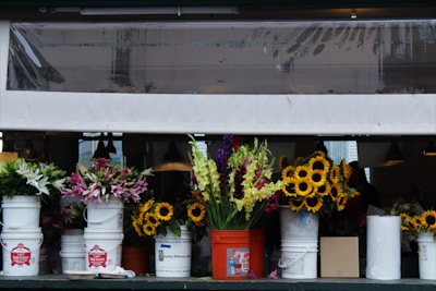 A row of colorful flowers arranged in white and orange buckets is placed on a shelf or counter. The flowers include sunflowers, lilies, and other assorted blooms in bright, vibrant colors. The scene is set indoors with dim lighting in the background and a protective plastic canopy partially visible above.