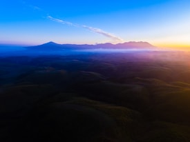 A breathtaking landscape with rolling hills bathed in the warm colors of a sunset. In the distance, majestic mountains are silhouetted against a vibrant sky transitioning from deep blue to orange and pink hues. Clouds softly float above the hills, adding depth and tranquility to the scene.