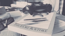 A black gaming controller rests on top of a book titled 'Theodore Roethke,' surrounded by books, a pair of eyeglasses, and newspapers on a cluttered surface.