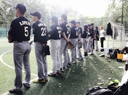Players in baseball uniforms stand in a line on a field, facing away. Most are wearing dark blue jerseys with numbers and names on the back. Hands holding gloves are visible, and various equipment is scattered around them. The field is surrounded by a fence, and trees can be seen in the background.
